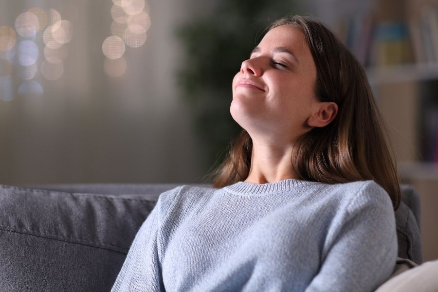 Frau sitzt entspannt auf einem Sofa mit geschlossenen Augen und geniesst einen Moment tiefer Ruhe und innerer Ausgeglichenheit